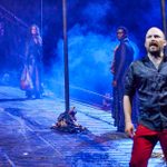 A dramatic stage scene with four actors. A man in red pants stands in the foreground with an intense expression. Three women in dark clothing are in the background on a smoke-filled wooden bridge. Blue lighting adds a mystical atmosphere.