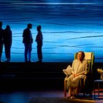 A person sits in an armchair in the foreground under a spotlight, looking contemplative. Behind, four silhouetted figures stand against an illuminated blue backdrop with abstract lines. A small table with a flower vase is next to the armchair.