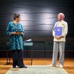 Two women stand facing each other on a stage. The woman on the left holds a tablet, wearing a blue patterned tunic and black pants. The woman on the right wears a chef's coat, striped trousers, a pink hijab, and holds a blue folder. A rug and table are in the background.