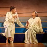 Two women sit on wooden steps, both wearing cream-coloured bathrobes. One holds a wine glass and smiles, while the other gestures with her hand. A bottle of wine and another glass are beside them. The backdrop is a warmly lit, minimalist setting.