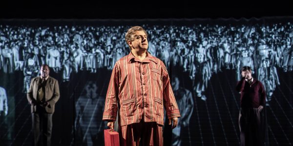 Michael Sheen wearing red striped pyjamas and holding a red briefcase. A large crowd of people are displayed on the screen behind him.