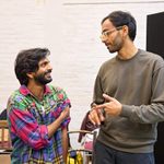 Two men are having a conversation indoors. The man on the left is wearing a colorful, patterned shirt, and the man on the right is wearing glasses and a grey sweater. They are standing in front of a white brick wall.