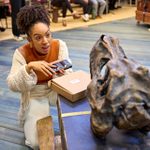 A woman kneels on the floor with excitement, holding a small metal tin and looking at a prop skull fossil. Behind her, multiple people are sat at the edge of the room.