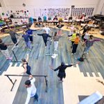 A large group of people practice ballet positions in a dance studio. They're standing with arms outstretched, using barres for support. Observers are seated at a large table in the background against a wall adorned with photos of costume and set designs. The floor is wooden and painted in a blue gradient.