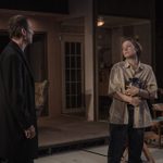 Two people are on a dimly lit stage. One person, wearing a coat, stands facing a person in a casual shirt holding an urn. They have an upset expression. Behind them is a dining table and chairs with wine and takeaway containers.