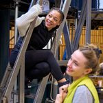 Two women interacting on a metal staircase. One woman in a black vest and white sweater sits on the steps, smiling and looking towards the other woman, who stands nearby in a yellow jacket. They are off to the side of a theatre set in a rehearsal room.