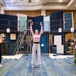 Three actors rehearse in a studio with stage props and numbered sections. The central actor has her arms raised in a ballet pose while the others stand to the sides. They are in a rehearsal room with a bare bones set behind them that has two levels and multiple doors.