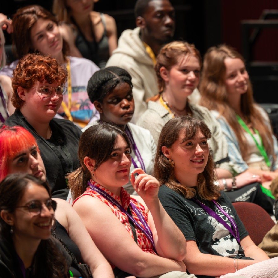 A group of diverse young people seated and attentively watching a presentation or performance. They are indoors in a theatre auditorium and appear engaged, with some wearing colorful lanyards or accessories.