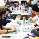 A group of people sitting at tables in a rehearsal room, engaged in arts and crafts. They are smiling and appear to be enjoying the activity. Various art supplies are scattered on the tables.