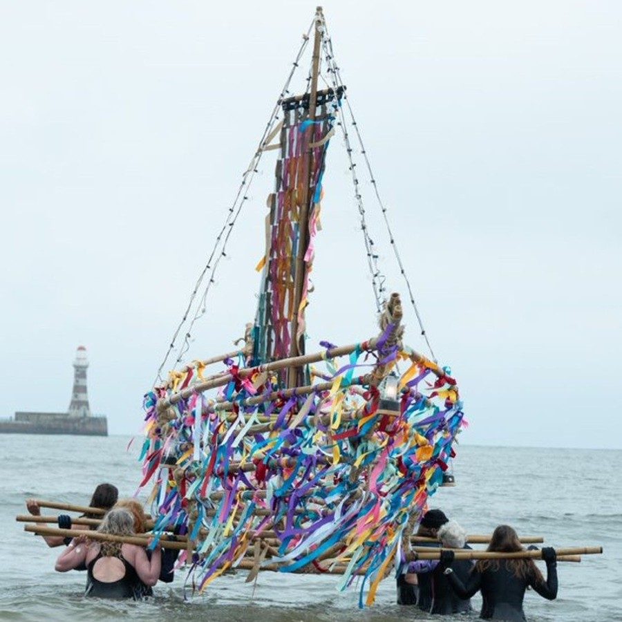 A wooden boat covered in ribbons being carried into the sea by six people