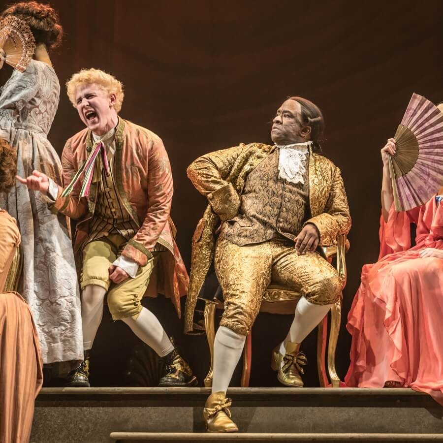 A theatrical scene features actors in ornate 18th-century costumes. One is passionately gesturing, while another sits confidently in a gold outfit. Others hold decorative fans against a dramatic backdrop.