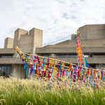 A wooden structure of a boat is covered in colorful, ribbons. It stands in tall grass against the backdrop of a the National Theatre building on a partly cloudy day.