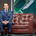 A man in a blue military uniform sits on a brown leather sofa. Behind him is a large map, partly visible. The room has a formal atmosphere.