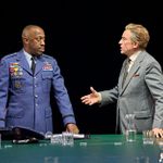 Two men are engaged in a discussion at a table. One is wearing a military uniform with medals and looks attentive, while the other, in a suit, gestures expressively. The setting is formal, with water pitchers and glasses on the table.