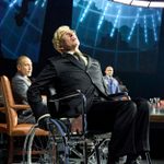 A man in a wheelchair, wearing a suit and glasses, leans back dramatically. Three men in suits sit at a table behind him, with a futuristic map display overhead. The setting suggests a formal or strategic meeting.