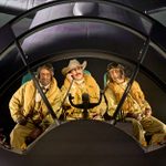 Three people in vintage pilot uniforms and goggles sit in an aircraft cockpit. One wears a cowboy hat. They appear to be in conversation or listening intently, surrounded by aviation controls and displays.