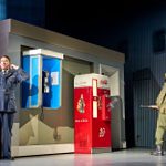 A stage scene features a man in a military uniform covering his ears in front of a phone booth and a Coca-Cola vending machine. Another man in army attire stands near the machine, holding a rifle. The setting evokes a mid-20th century vibe.