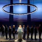 A theatrical stage scene with a woman in a long, light-colored dress standing in the center under a circular, illuminated fixture. She is surrounded by a group of people dressed in dark clothing, lined up on either side. The backdrop displays towering structures.