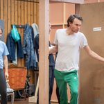 A man in green sweatpants and a white t-shirt opens a door. A woman in a blue t-shirt stands nearby, smiling. The room has wooden walls, furniture, and clothing hanging in the background.