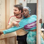 Two people lean playfully around a doorframe. The man wears a t-shirt, and the woman wears a teal sweatshirt. They both are looking at something off-camera.