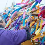 A person wearing a purple coat and glasses ties colorful ribbons onto a bamboo structure. The ribbons, including red, blue, yellow, and pink, create a vibrant display. The person's hair is gray, and the background is blurred.