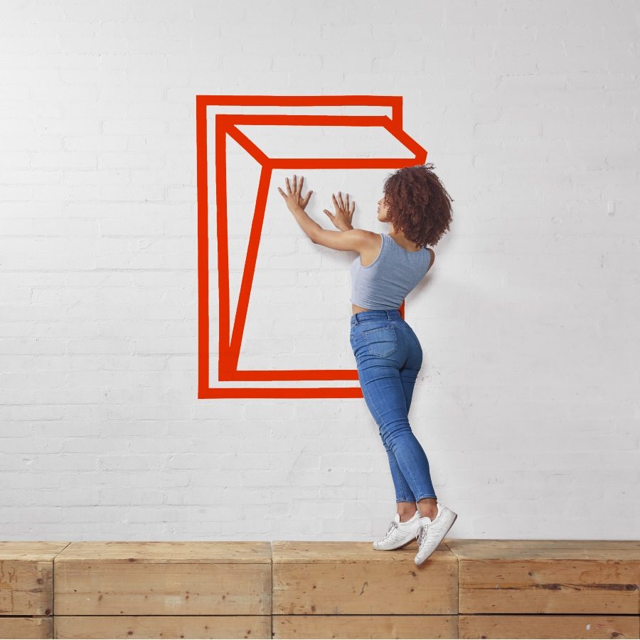 A person in a grey t-shirt and sweatpants poses inside a red 3D cube outline painted on a white brick wall. The person appears to be interacting with the cube's structure.