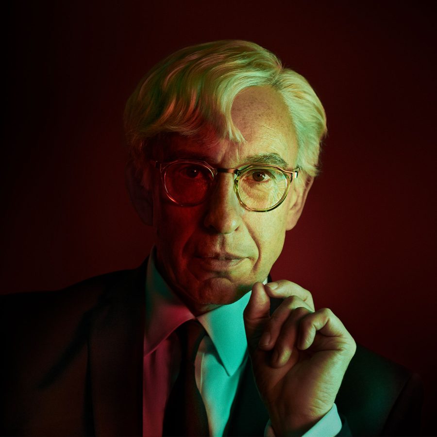 A photo of a man wearing glasses and a suit, with his hand near his chin, set against a dramatic red and green gradient background. His expression is thoughtful and serious.