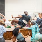 A group of people are gathered in a studio, striking poses around a man sitting on a piano.