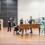 A group of performers rehearse around a grand piano in a spacious studio. Some kneel, others stand or sit, striking various poses. The setting includes wooden floors and high ceilings.