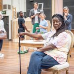 A woman with a cane sits on a chair in a rehearsal studio while others stand and watch.