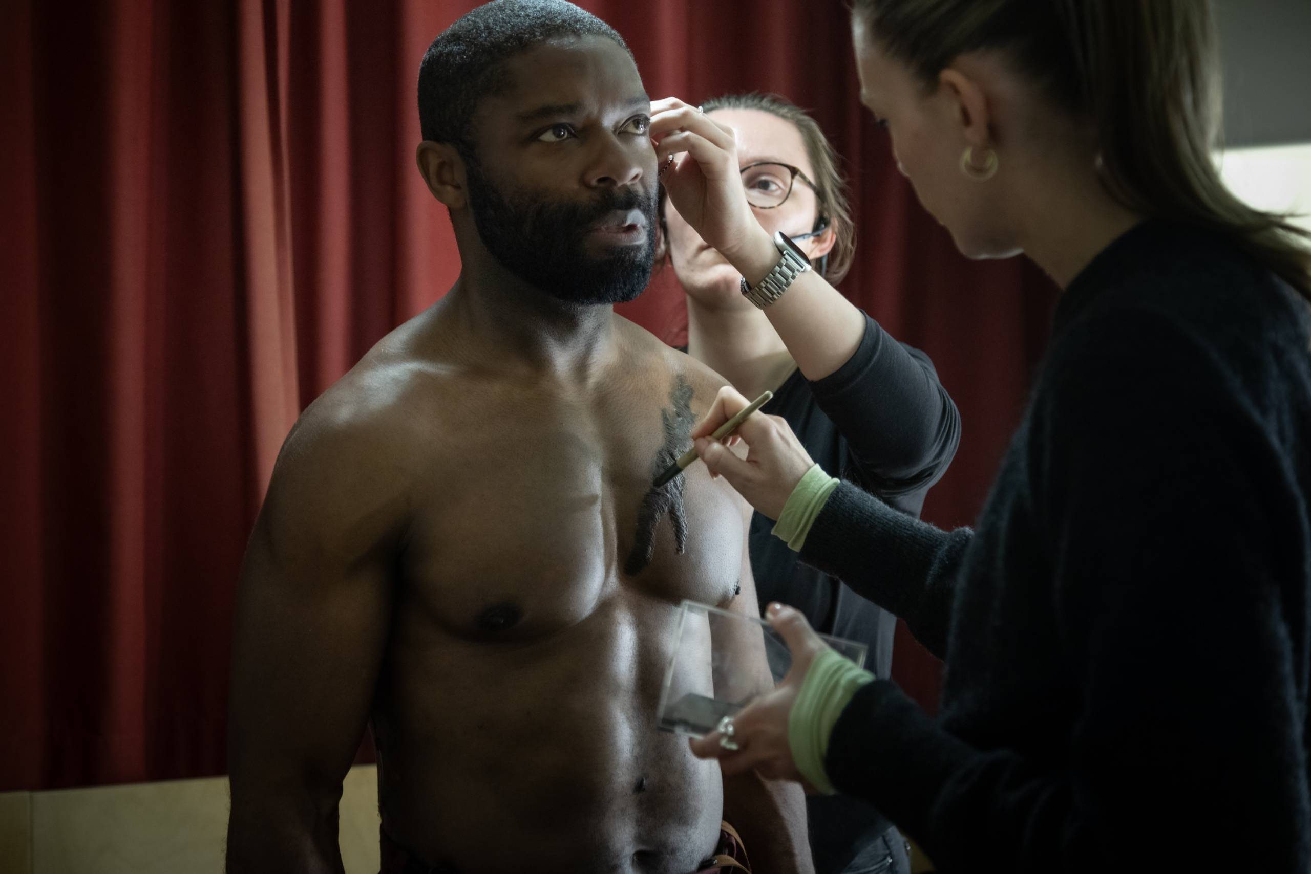 A shirtless man with a beard is having makeup applied to his face and chest by two makeup artists. One is applying makeup near his eye, and the other is working on his chest. A red curtain is in the background.