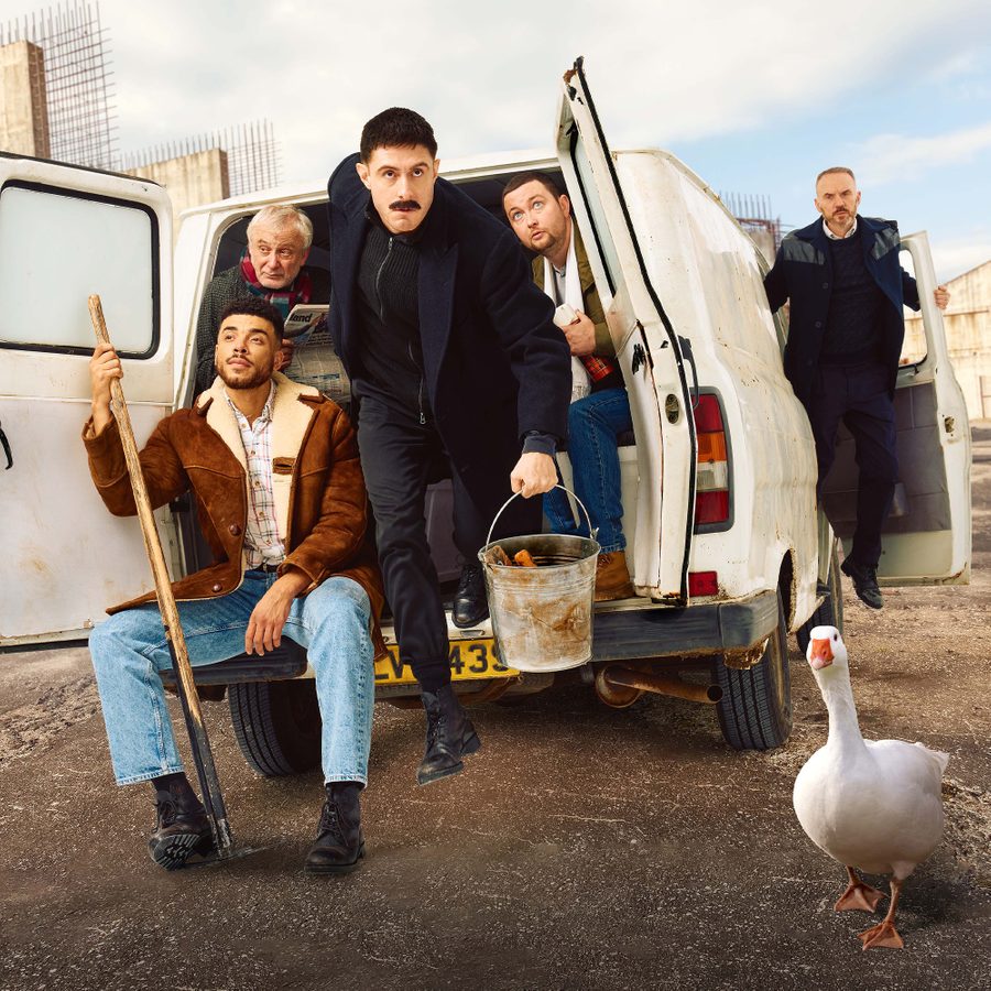 A group of five individuals pose around and inside a white van in an open concrete area. One person holds a mop and bucket, another sits with a shovel. A goose walks in the foreground.