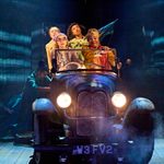 Four actors in vintage attire are sitting in a classic car on stage. The headlights are on, and there is a smoky effect around them. Another cast member is visible in the back to help move the car around the stage.