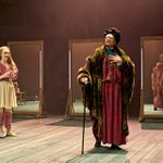 Two actors on stage in a theatrical production with four large mirrors behind them. A young actress wears a pink cardigan and legwarmers, and a white skirt. She is holding a pair of ballet shoes. The other actor is dressed in a fur coat with a cane, standing confidently with a hand on their chest.