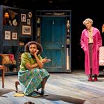 Two actors perform on stage. One sits on a small stool with an expressive pose, wearing a green jumper and floral skirt. The other stands nearby in a pink robe, looking towards them. The set includes a blue door, shelves, and various photographs and decorations.