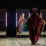 An actor dressed in a lavish, vintage-style fur coat stands facing three large mirrors, reflecting their image. Projected into the mirror in front of them is the actor playing a young version of their character, wearing the same headwrap and a similar dress.