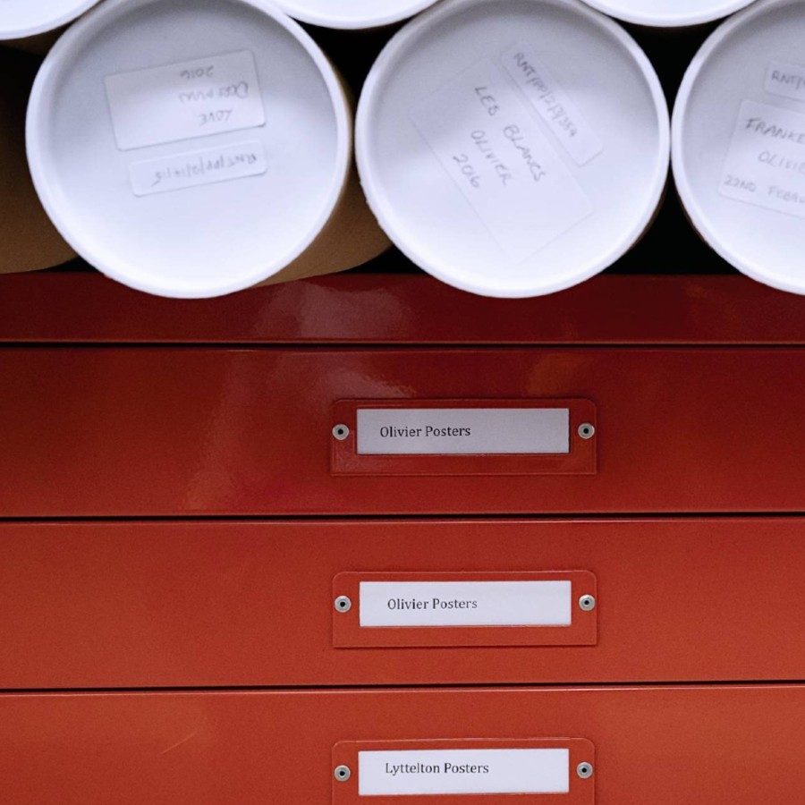 A row of labeled poster tubes is stacked on a red filing cabinet.