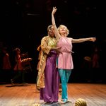 Two performers on stage, one in a gold and purple outfit supporting a dancer in a pink and blue outfit. The dancer extends an arm, balancing on one foot. The background shows other performers moving pieces of furniture.