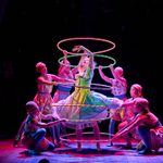 A group of performers in colourful costumes surrounds a central dancer with neon hula hoops. The scene is set on a dimly lit stage, with vibrant lights focusing on the dancers.