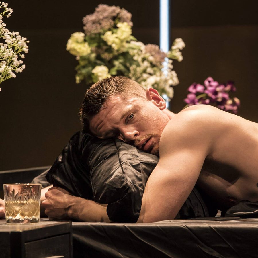 A shirtless man with a tattoo lies on a bed, resting his head on a pillow. The setting is adorned with elegant flower arrangements in vases and a glass candle holder, creating a serene atmosphere.