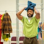 An actor in a rehearsal room wearing a lime green jumper. He has a pained expression and is holding a pair of blue trousers to his head. Behind him are railings with colourful clothes hanging from them.