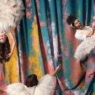 Three performers in whimsical costumes pose against a colorful, painted backdrop. One is upside down on a trapeze, another stands holding a prop, and the third hangs from aerial hoops. They wear fluffy dresses and expressive accessories.