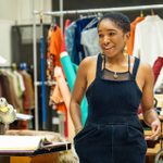 An actor in a rehearsal room wearing a blue jumpsuit and yellow jewellery. On her left is a sewing machine and an open script. Behind her, colourful clothes hang from a railing.