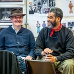 Two actors in a rehearsal room, sat on chairs. On the wall behind them are printed out images of the set design, costume designs and references.