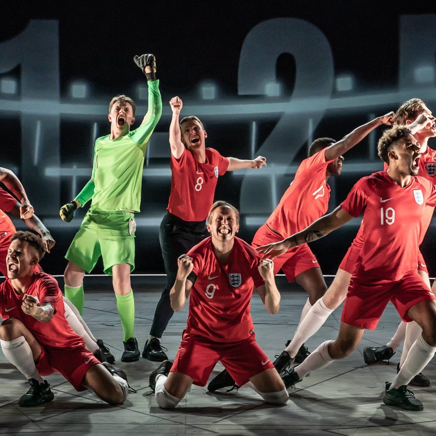 A group of men are in a group, they all wear red England kit and are in various stages of celebration after a goal. Some are on their knees, all are exclaiming. One of the men is in a green goalie uniform.