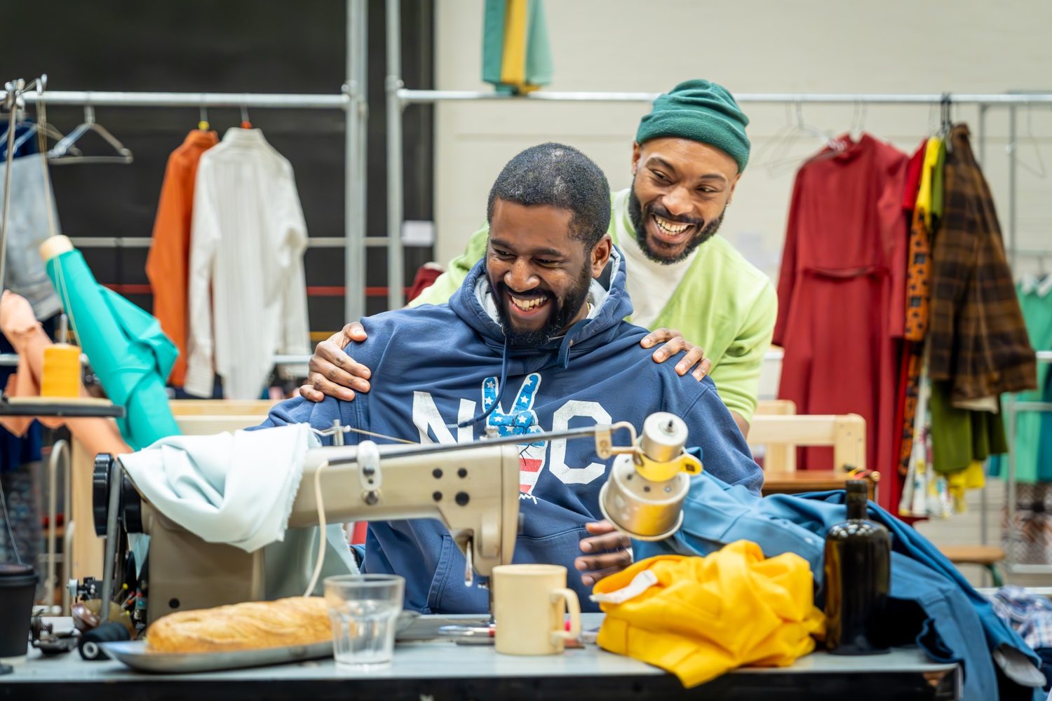 Two actors in a rehearsal room laughing. One is sat at a table with a sewing machine and piles of clothes. The other actor is stood behind him and holding his shoulders.