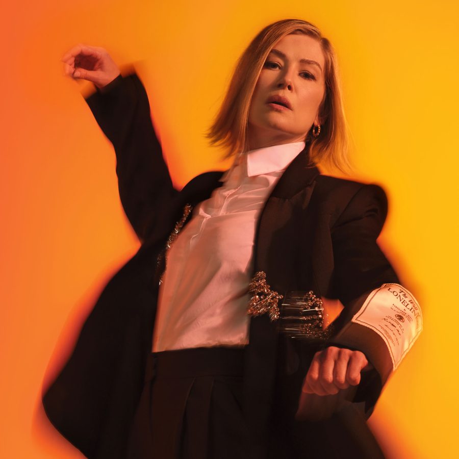 A person with shoulder-length hair poses in a dynamic stance against an orange background. They wear a black blazer with gold embroidery over a white shirt. One arm is raised, and the other has a fabric band with text.