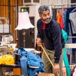 An actor in a blue jumper and red neckerchief using a broom. He is stood next to a table with a sewing machine and a pile of clothes. Behind him is a railing with colourful clothes hanging from it.