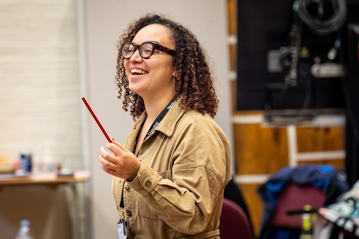 A woman in a tan jumpsuit and tortoiseshell glasses looking to the side and laughing. She is wearing a lanyard and holding a pencil.