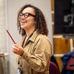 A woman in a tan jumpsuit and tortoiseshell glasses looking to the side and laughing. She is wearing a lanyard and holding a pencil.